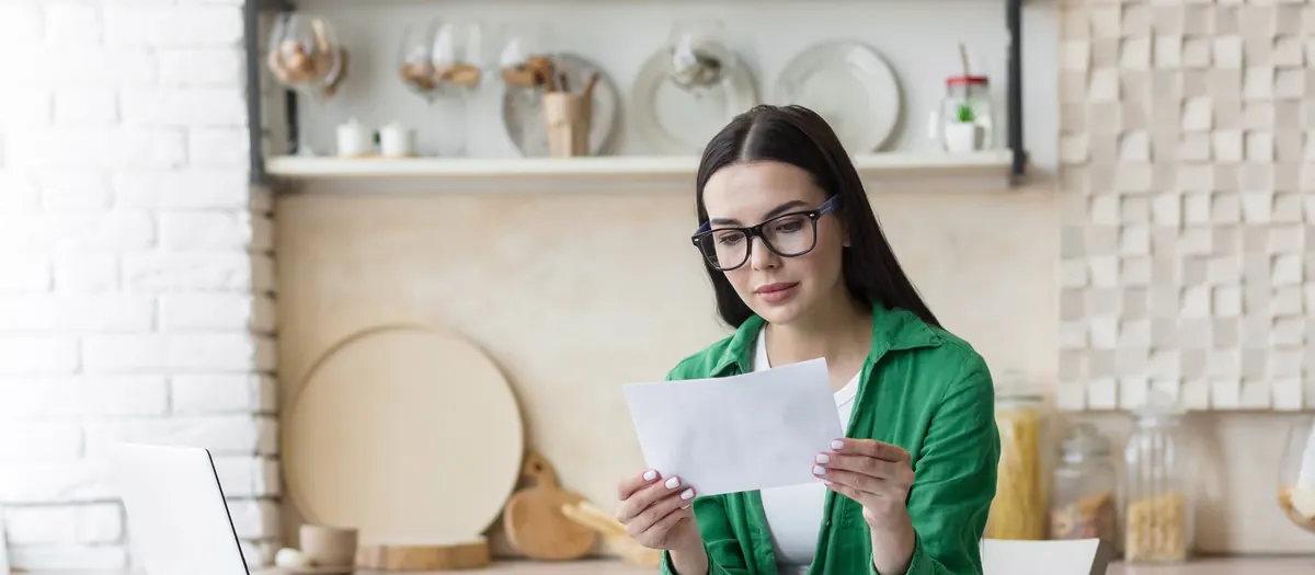A woman at a kitchen counter reading a letter, a laptop and papers beside her.