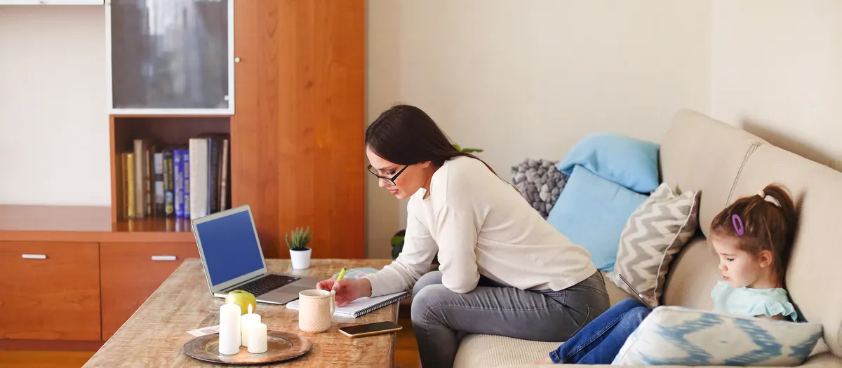 A woman working at a laptop on a coffee table at home, a young child sitting on the sofa beside her.